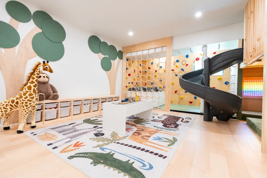 A children's playroom interior with a spiral slide, climbing wall, tree wall decals, a giraffe plush toy, and a large animal-themed rug on a light wood floor.