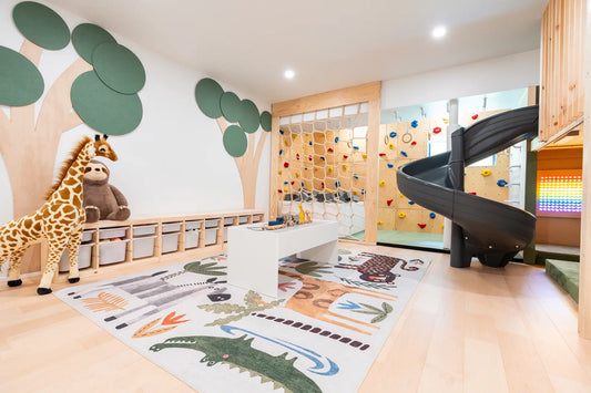 A children's playroom interior with a spiral slide, climbing wall, tree wall decals, a giraffe plush toy, and a large animal-themed rug on a light wood floor.