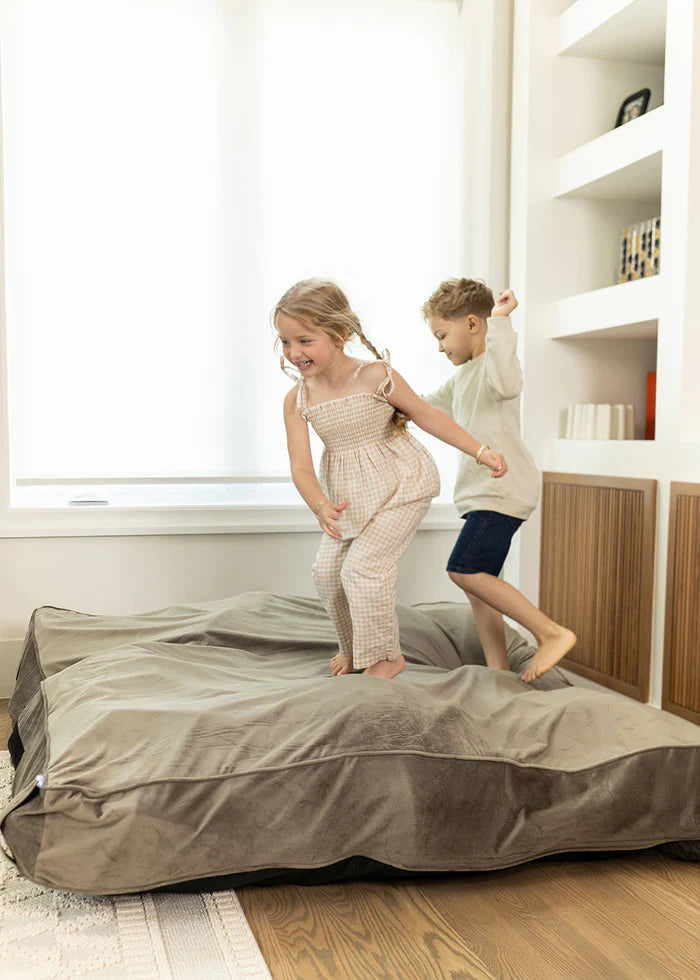 Two children playing on a large bean bag in a room with shelves and a window.